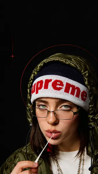 Billie Eilish trägt eine olivgrüne Kapuzenjacke und eine marineblau-weiße Supreme-Mütze, während sie Kaugummi kaut